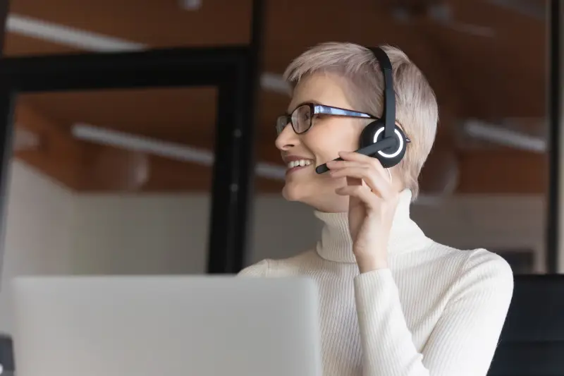 Happy young short haired 30s businesswoman in eyeglasses and headset with microphone consulting with colleague about decision, discussing contract or working issues with client online in office.